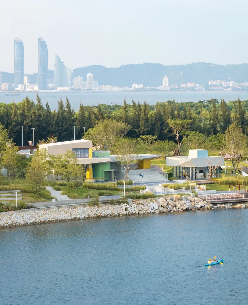 Distant view of waterfront pavilions nestled in parkland with kayakers on the water