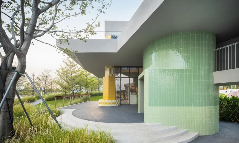 Covered entry with green tiled cylindrical volume and yellow pixelated column under deep concrete overhang