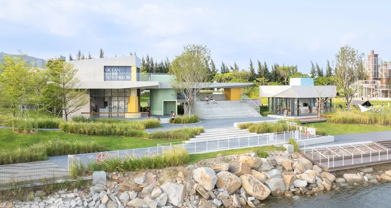 Waterfront complex with colored volumes and native grasses along a rock-edged shore in daylight