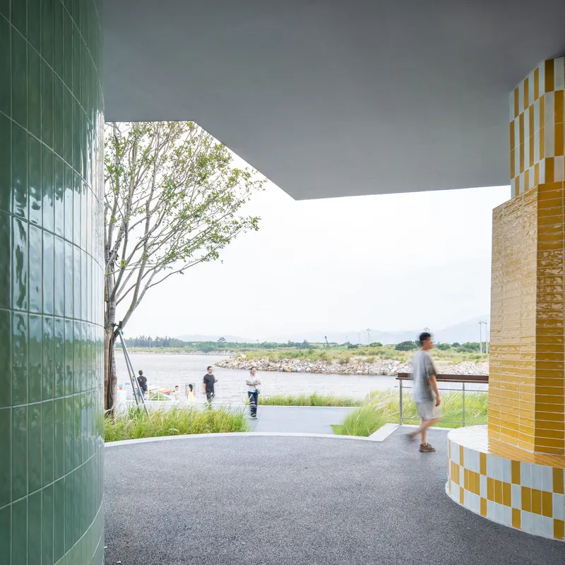 Covered walkway framed by green tiled wall and yellow mosaic column opening to waterfront grasses