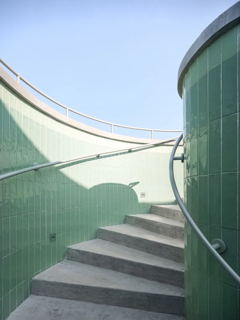 Curved concrete staircase with metal handrail ascending between green glass block walls under open sky