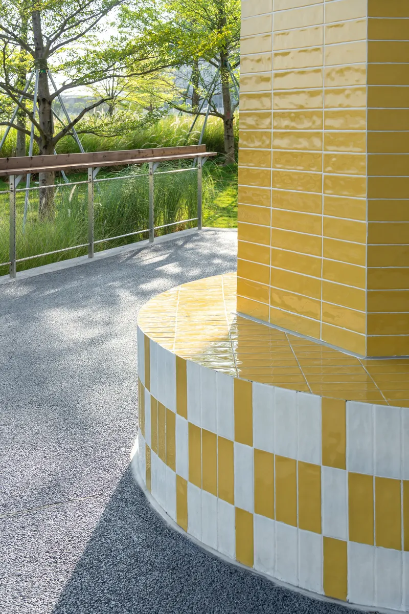 Curved yellow and white checkered tile wall meeting a gravel path with stainless steel railing and trees beyond
