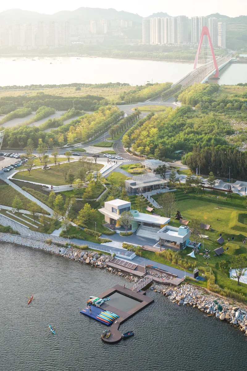 Aerial view of white pavilion complex near waterfront with red suspension bridge and forested hillside beyond