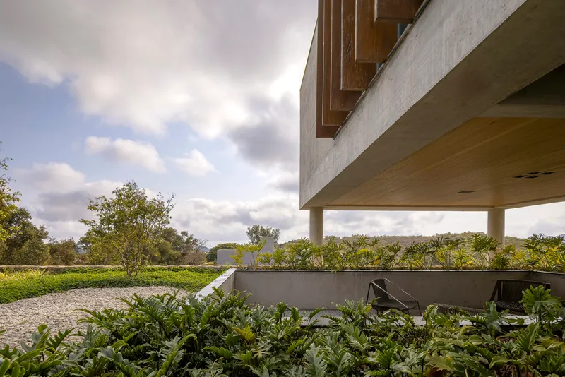 Cantilevered concrete volume above a planted terrace with gravel path and cloudy sky