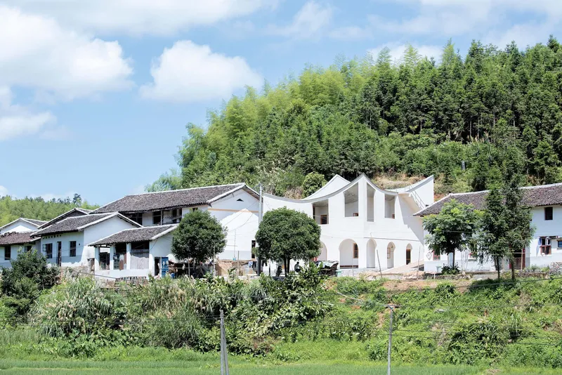 White arched facade rising between traditional tile-roofed houses with cultivated fields and bamboo forest behind