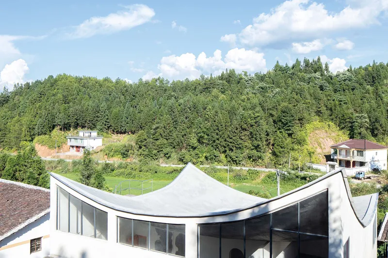 Curving white roof canopy above floor to ceiling glazing overlooking the valley under summer clouds