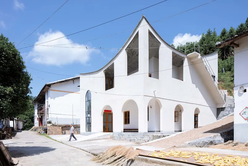 White facade with arched openings and a curved gable roof fronting a village street under construction