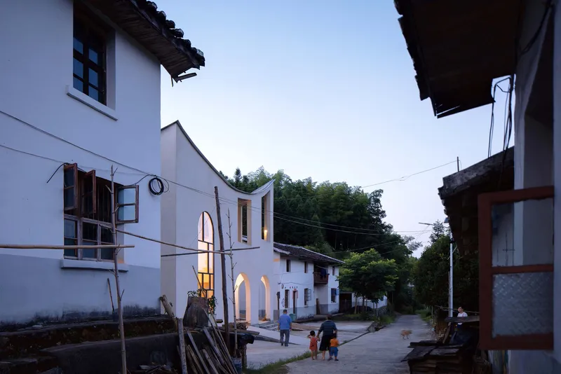 Street view at dusk with residents walking past the illuminated arched entrance between neighboring village houses