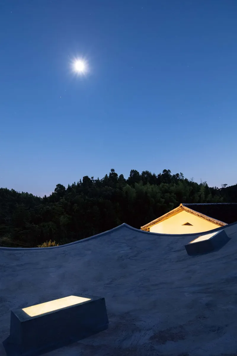 Rooftop terrace with curved parapets and a lit gable skylight under moonlight against a forested hillside