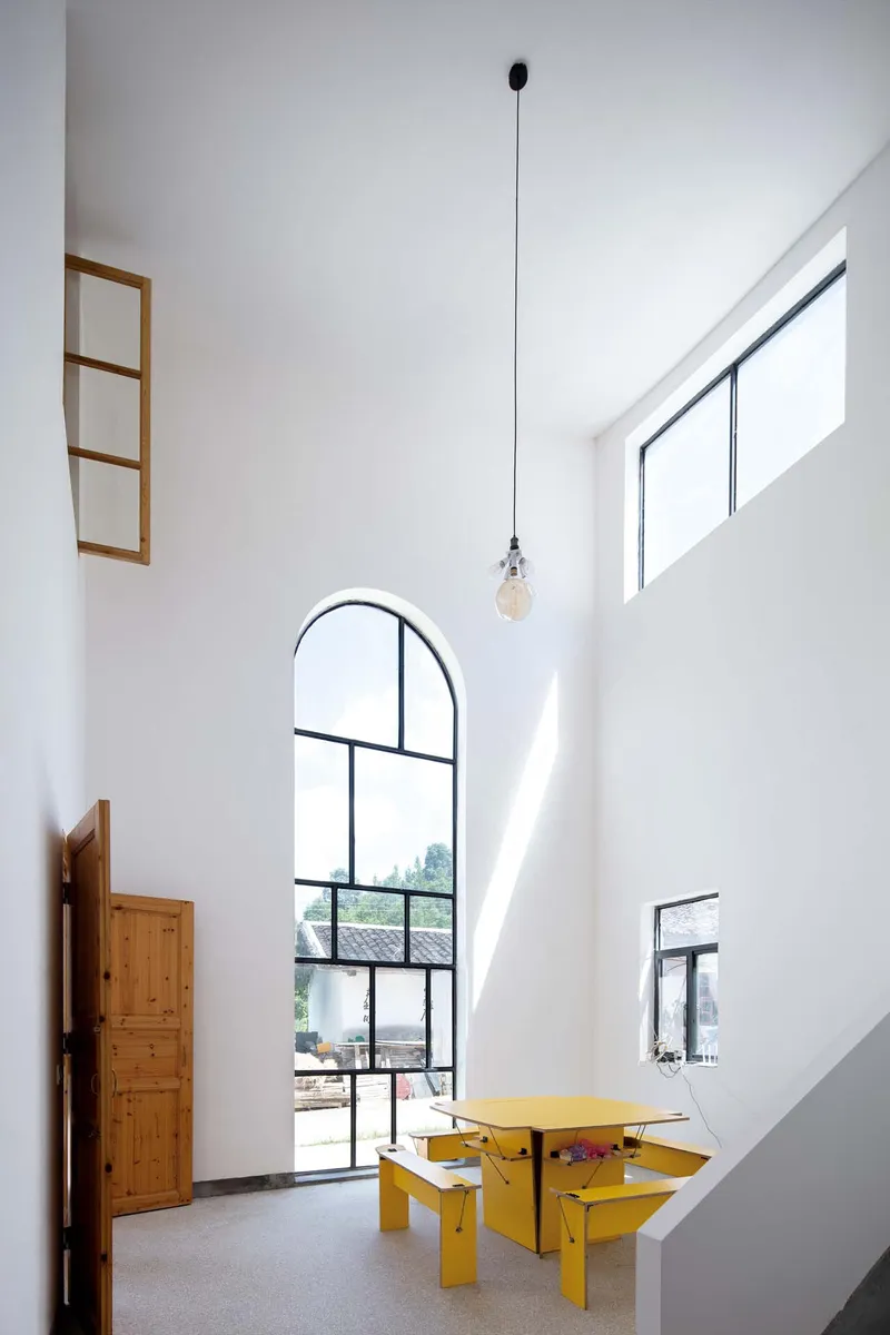 Tall arched steel window framing trees outside, with yellow table and pendant bulb in white interior