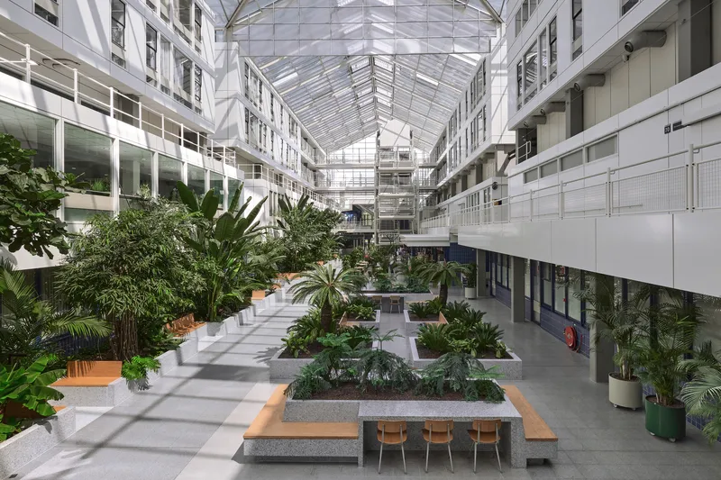 Multi-story interior atrium with glass roof, planted beds, and timber benches along a tiled central corridor