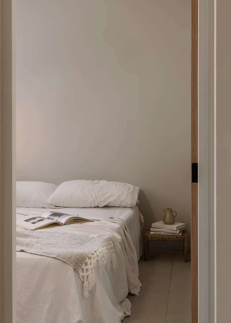 View through doorway into bedroom with unmade bed and books on woven stool bedside table