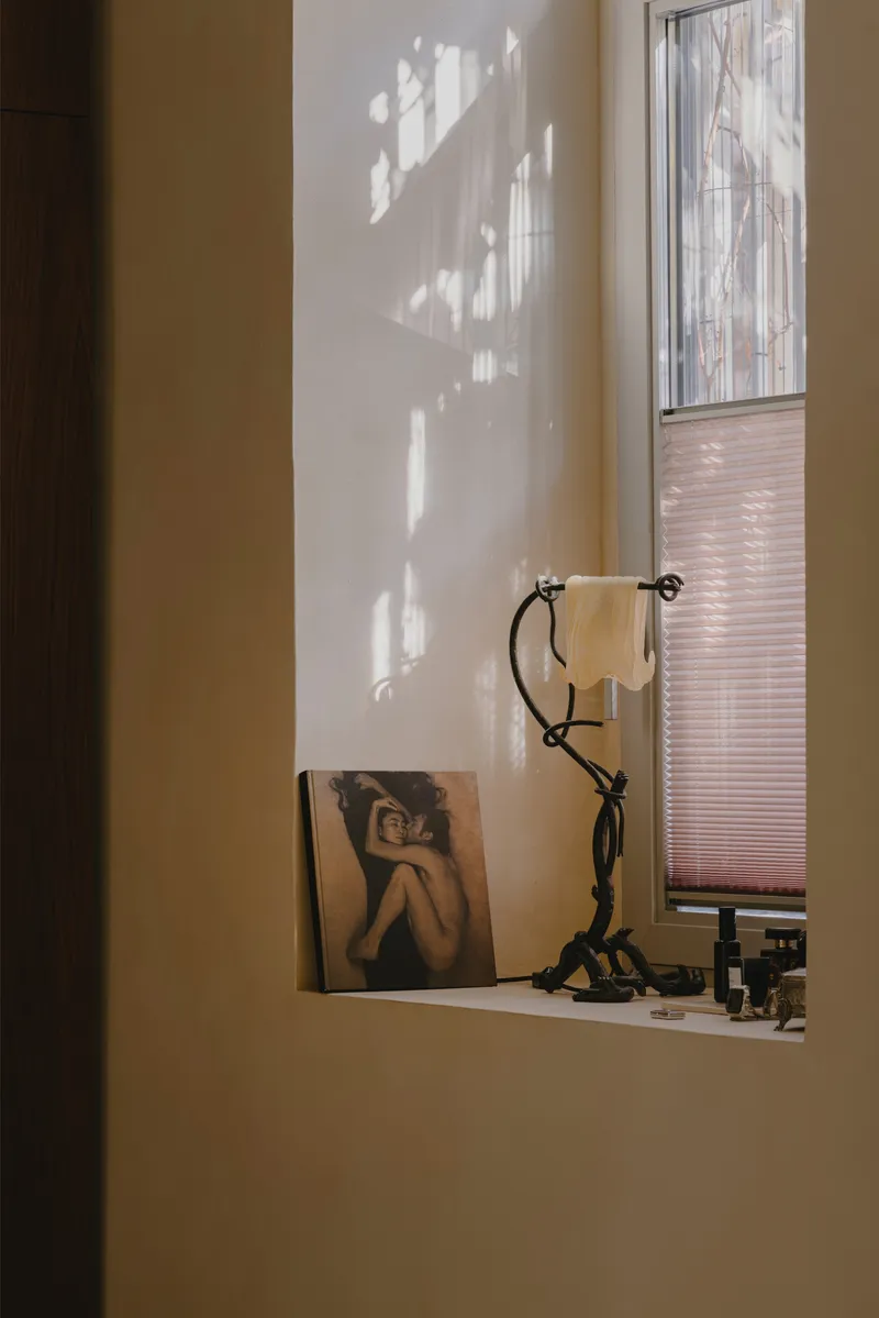 Shallow wall niche with a sculptural lamp and artwork framed by dappled afternoon light through a pleated shade