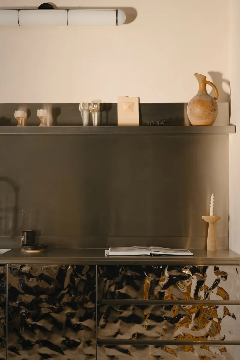 Kitchen counter with brushed bronze backsplash and sculptured metallic cabinet fronts beneath floating shelves at dusk