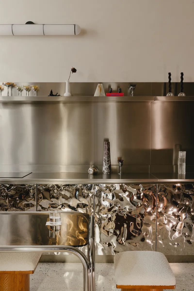 Kitchen island detail with reflective crumpled metal cabinetry and terrazzo countertop below stainless steel shelving