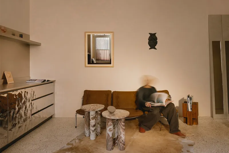 Living room with terrazzo flooring, tan sofa, carved timber side tables, and a person in motion