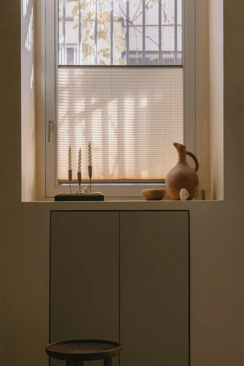 Window sill with pleated shade and terracotta jug catching afternoon sunlight above minimal cabinetry
