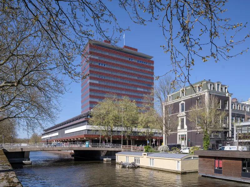 Horizontal brick and glass tower rising above a canal with houseboats and spring tree branches