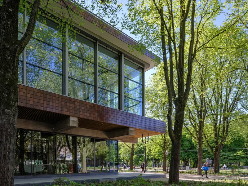Cantilevered brick and glass volume on timber columns above a tree-lined park path with pedestrians and cyclists