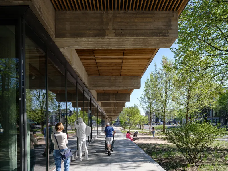 Cantilevered concrete overhang with timber soffit shading a glazed corridor where visitors walk beneath spring trees