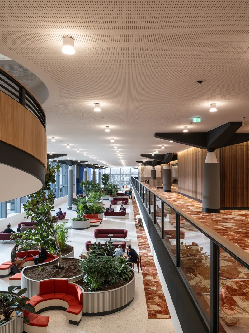 Multi-level atrium with circular planters, red seating, and terrazzo flooring under a perforated acoustic ceiling