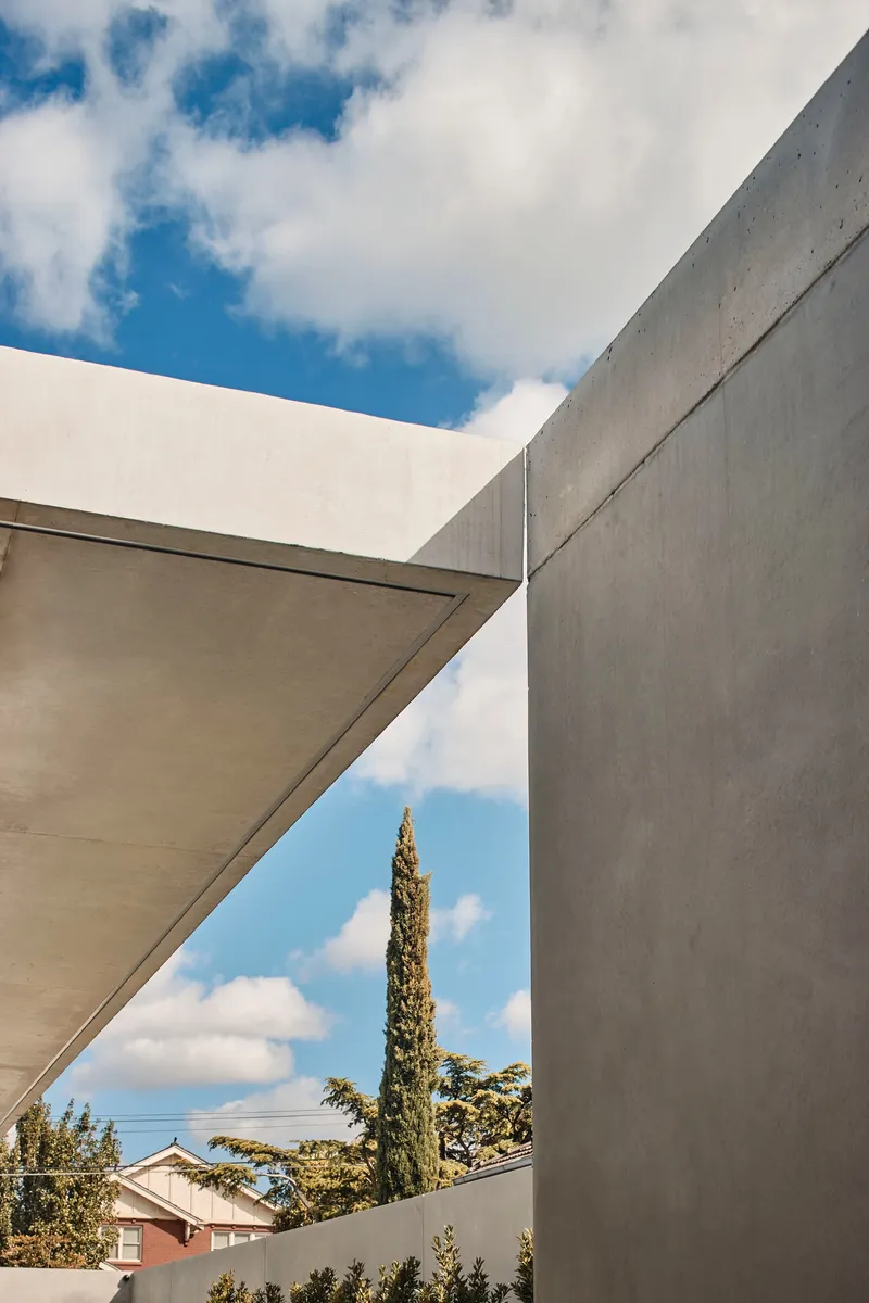 Upward view between concrete volumes and a cantilevered overhang framing a cypress in blue sky
