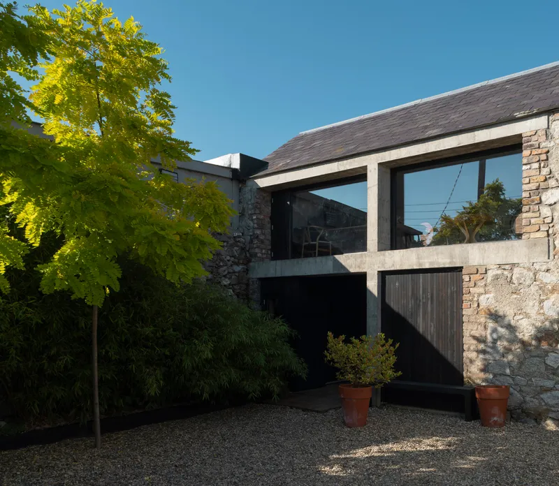 Slate-roofed extension with concrete frame and timber cladding beside a stone wall and potted trees