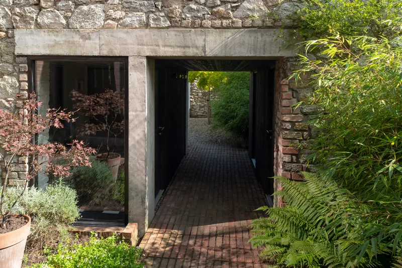 Concrete portal framing a brick-paved passage through a stone wall with planted beds and ferns