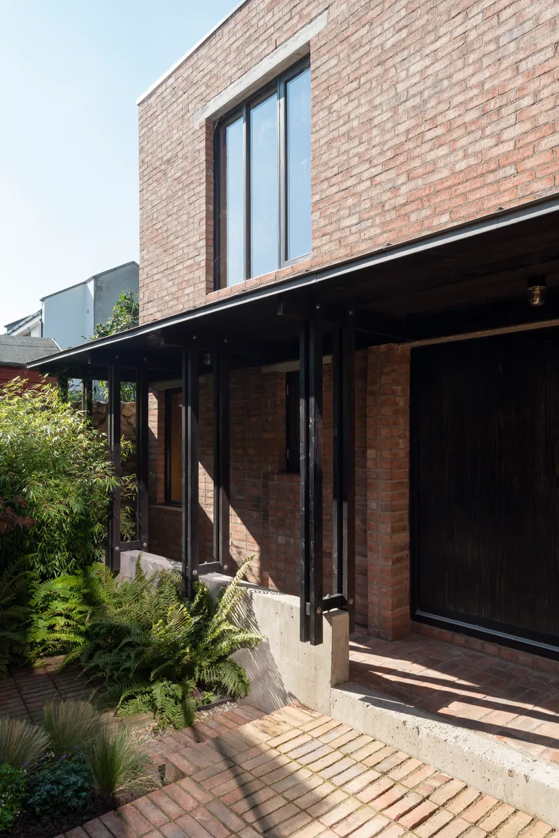 Corner of brick volume with black-framed window above cantilevered roof sheltering planted courtyard with ferns