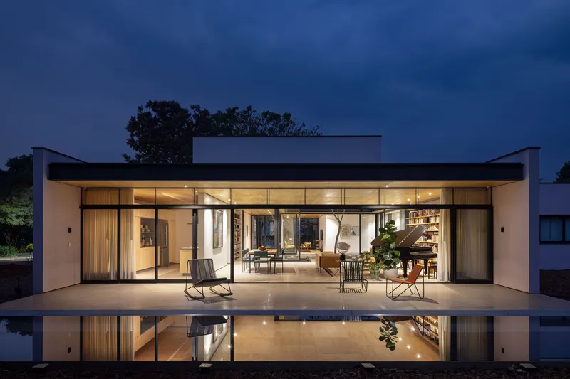 Fully glazed facade opening to terrace and reflecting pool with interior furniture visible at twilight