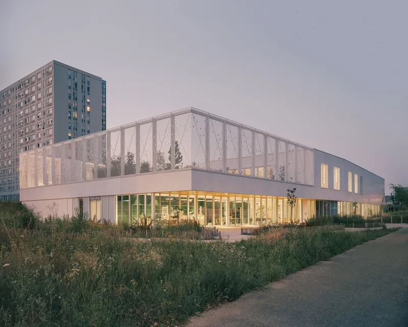 Corner view of the metal mesh-wrapped facade at twilight with residential tower behind and grasses in foreground