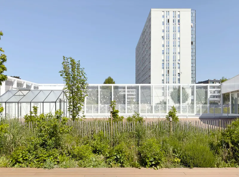 Perforated white metal screen facade with greenhouse and planted beds in the foreground