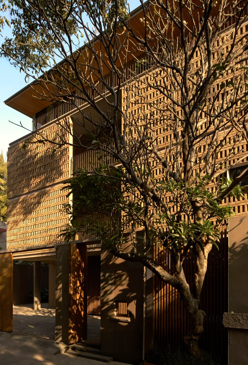 Layered perforated brick facade with deep overhangs behind a bare winter tree in afternoon light
