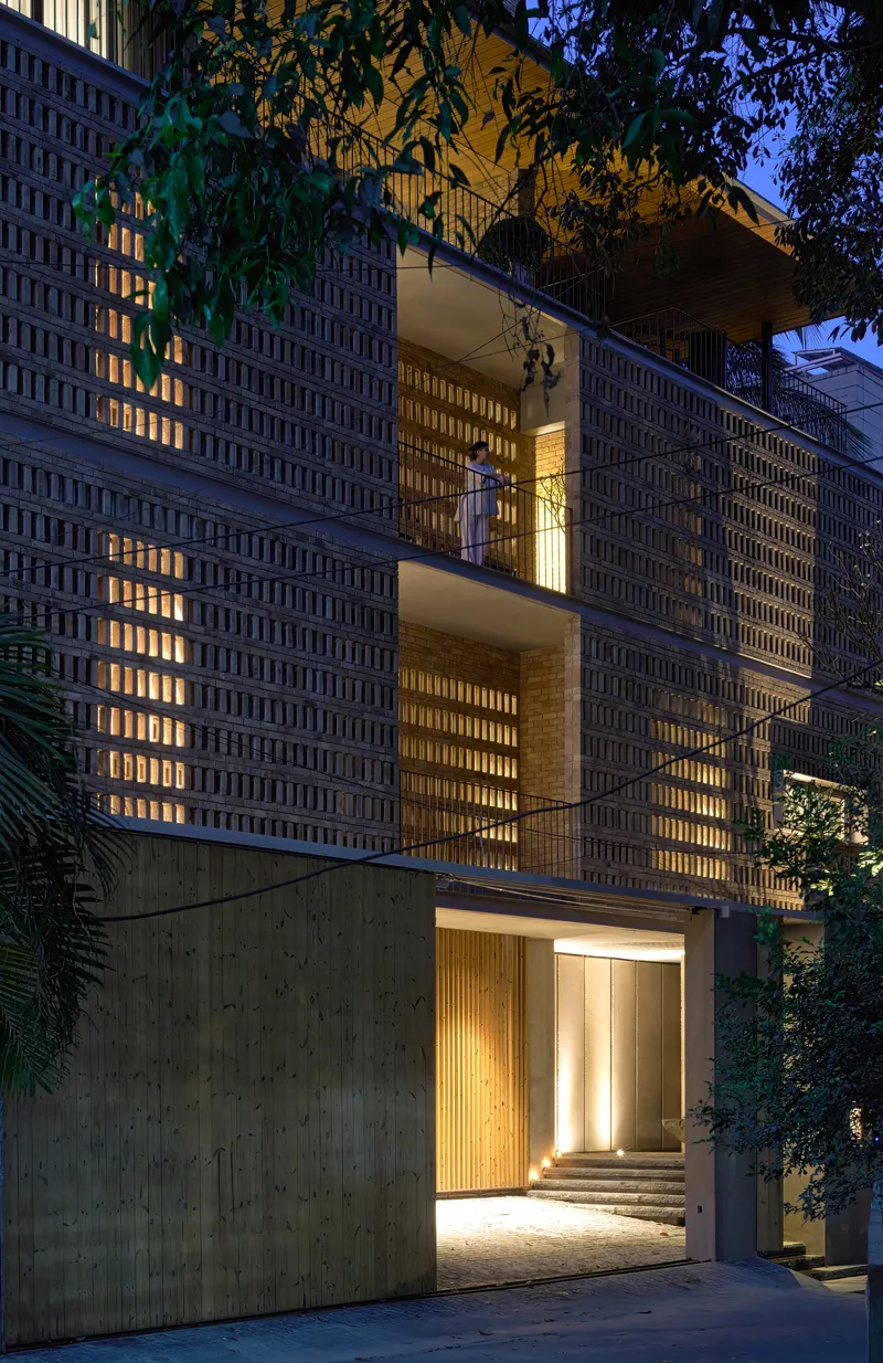 Illuminated perforated brick facade with stacked balconies and entrance at dusk surrounded by vegetation