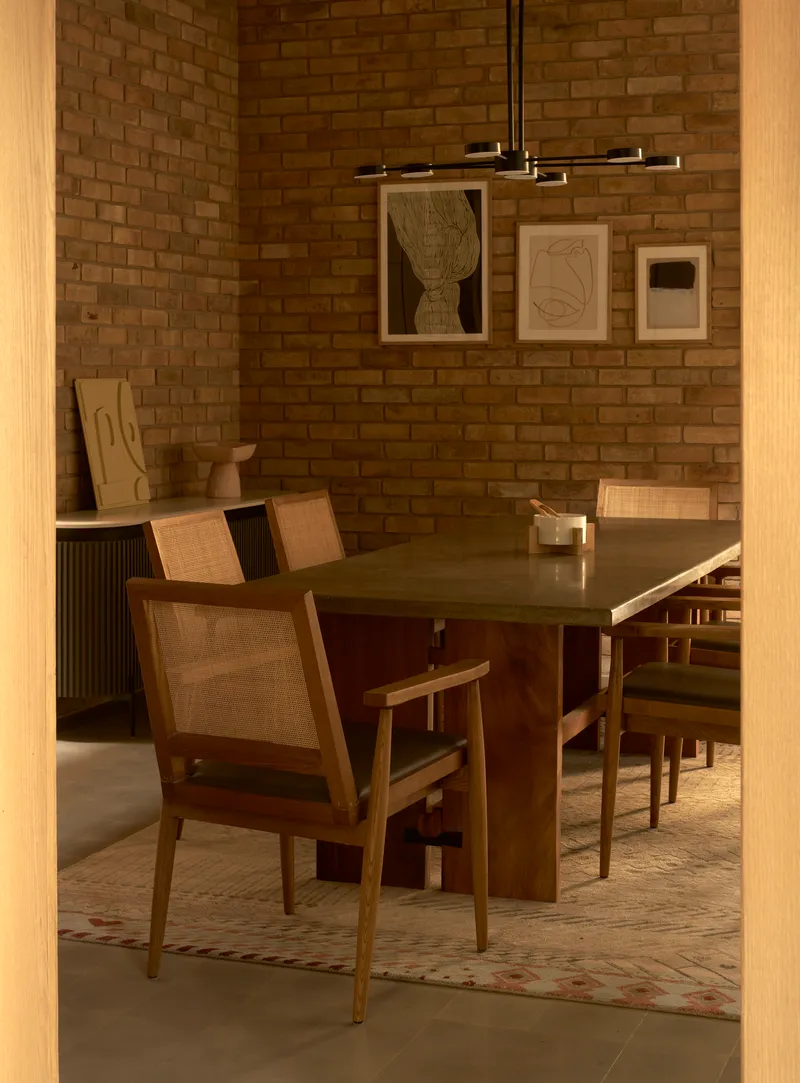 Dining room with brick wall, timber table and chairs, and framed artworks lit by pendant fixture