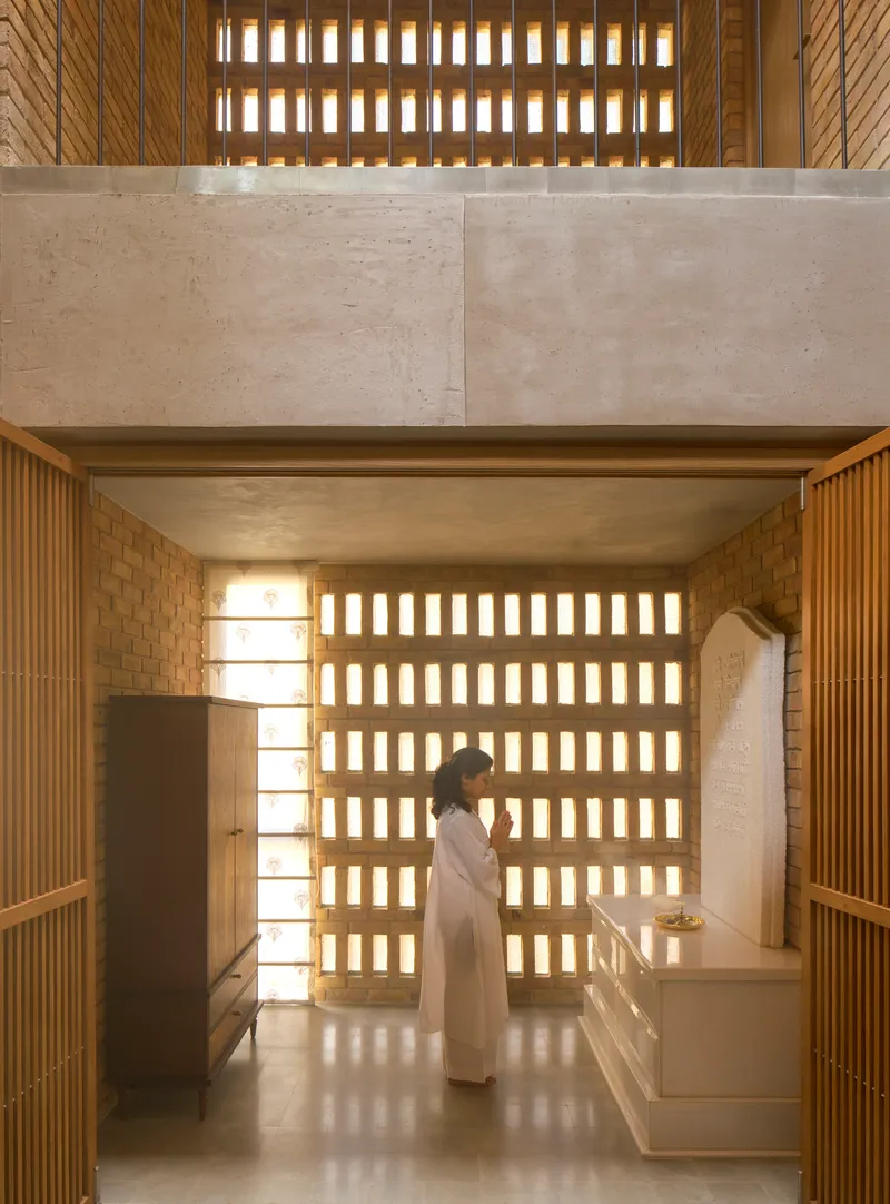 Double-height kitchen with gridded brick screen wall and figure standing at counter beneath concrete mezzanine
