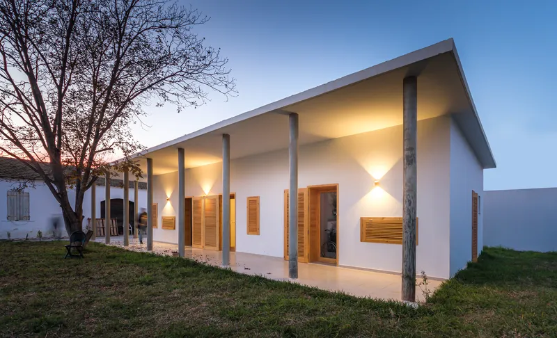 White facade with timber louvered openings and columned porch illuminated at twilight beneath a bare tree