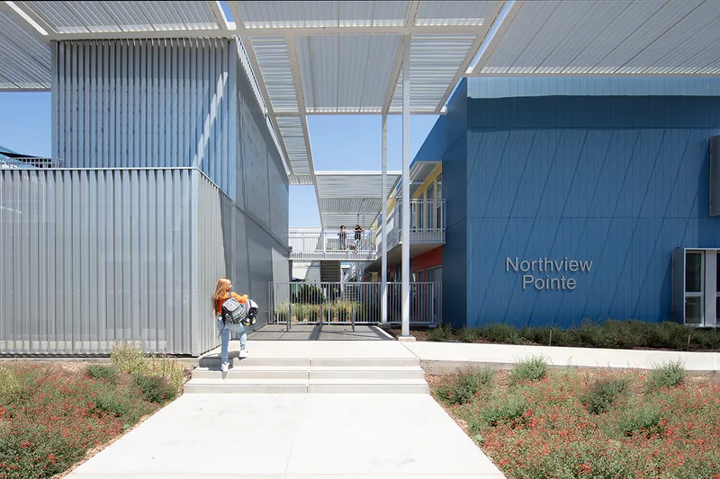 Central courtyard walkway with corrugated metal volumes and white canopy overhead under clear skies