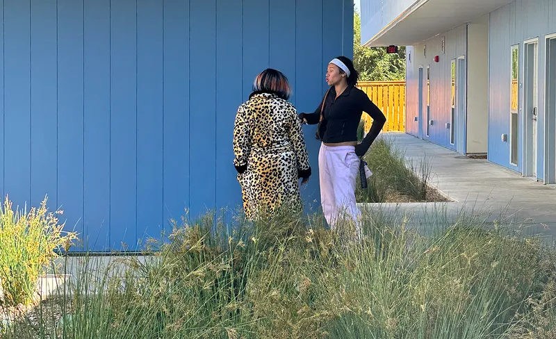 Two people conversing beside the blue panel facade and landscape buffer at the covered walkway edge