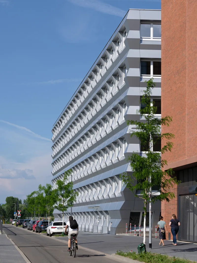 Angled facade with projecting white shading elements and a cyclist passing on the street