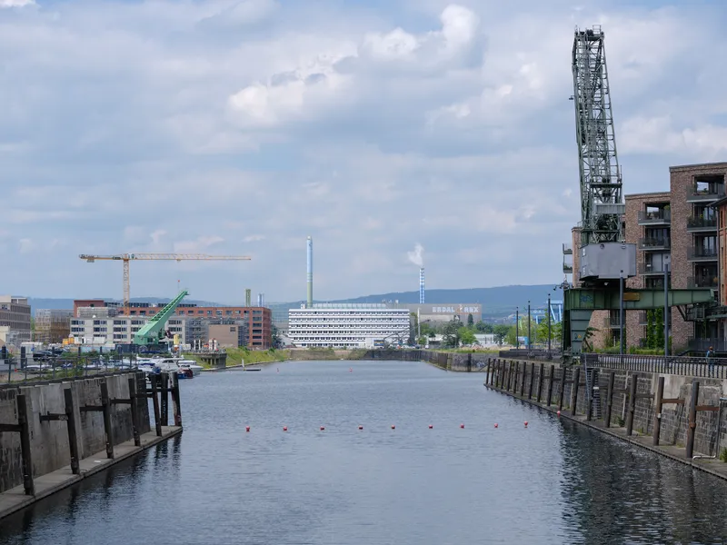 View across the canal showing the layered white facade with a crane and industrial chimney under scattered clouds