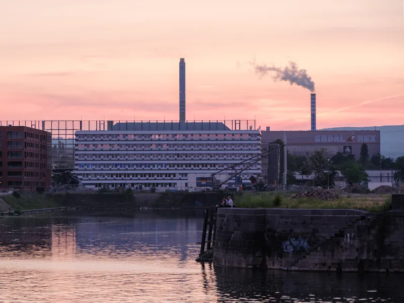 Horizontal facade reflected in the canal at dusk with a distant smokestack visible against the pink sky