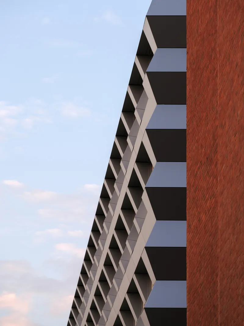 Diagonal facade with white sunshading elements meeting the vertical red brick tower volume at dusk