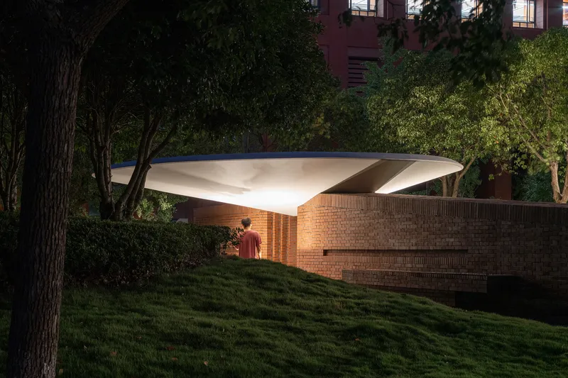 Illuminated canopy sheltering a brick pavilion wall as a figure stands on the lawn at twilight