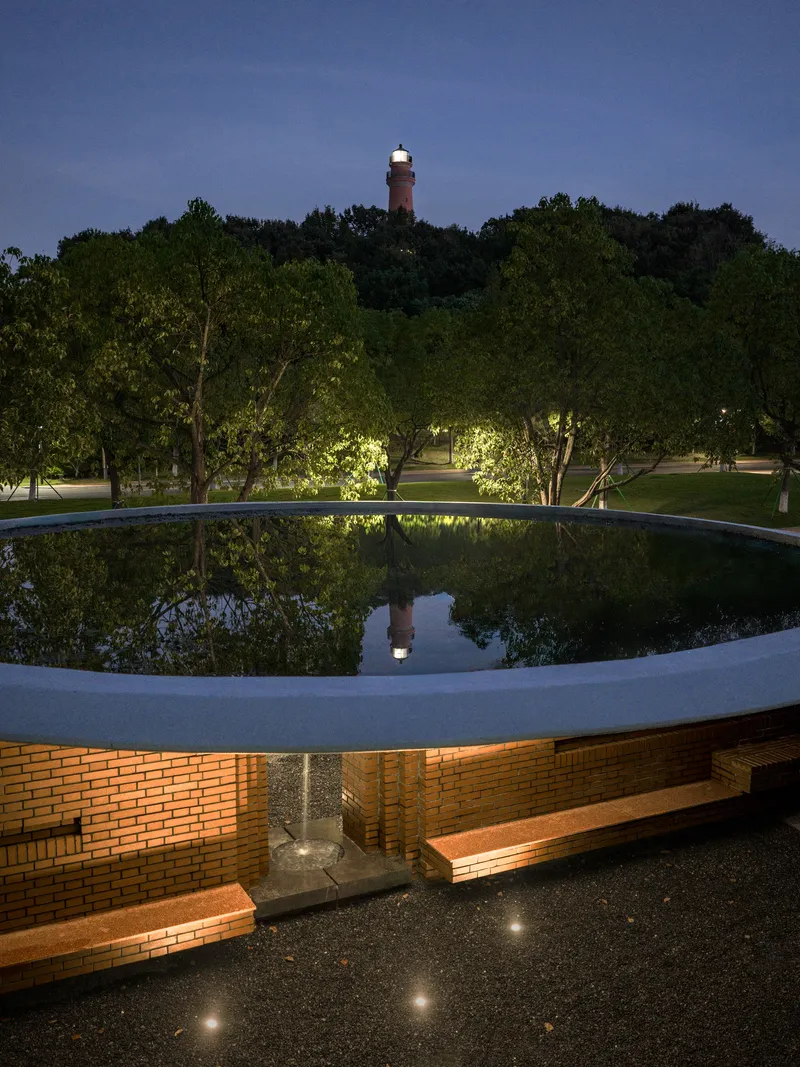Circular reflecting pool atop a brick pavilion at night with lighthouse glowing in the distance