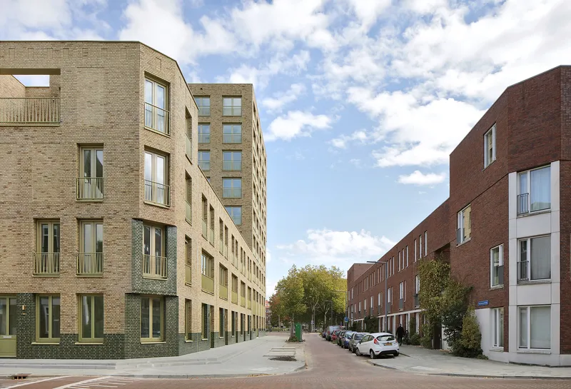 Residential street lined with buff brick housing blocks and parked cars under scattered clouds