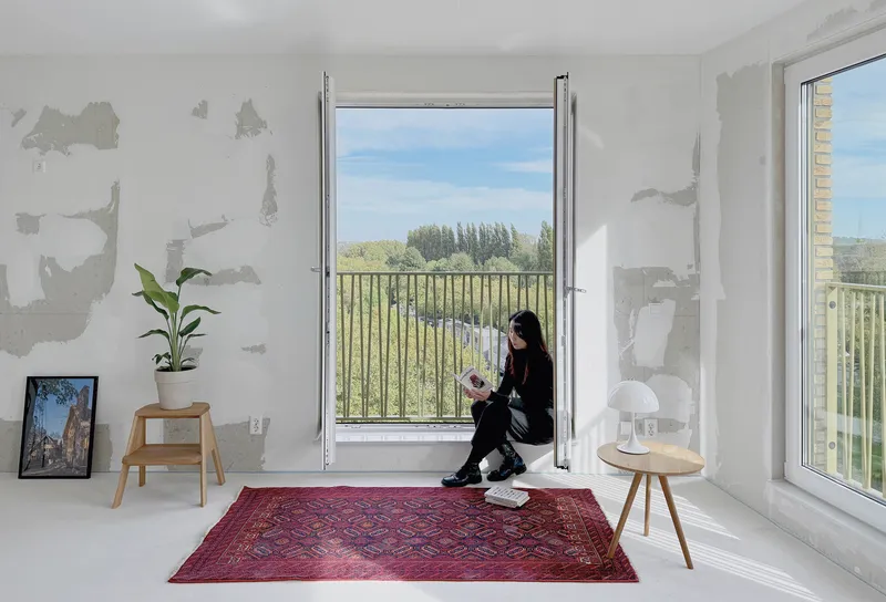 Person seated in the window threshold reading as sunlight streams across unfinished plaster walls