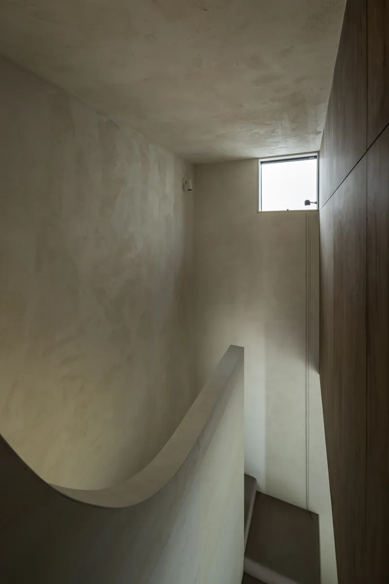 Curved plaster stairwell with dark timber wall and narrow horizontal window casting daylight