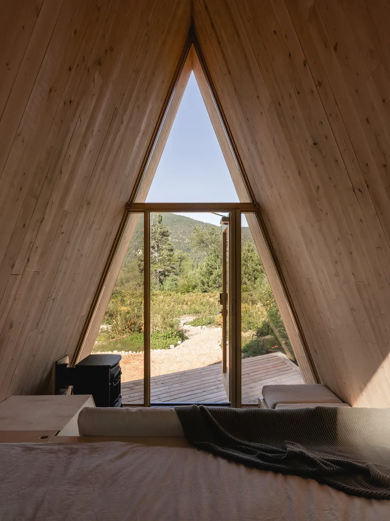 Interior view through triangular glazed opening framing timber deck and mountain landscape beyond a bed