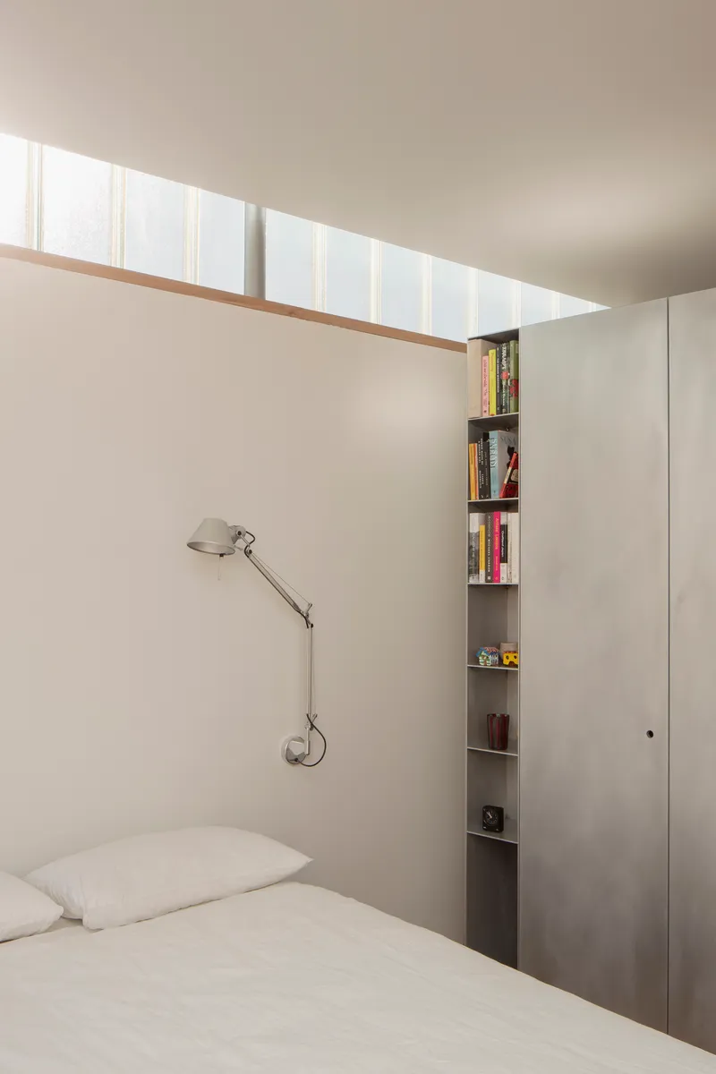 Bedroom with clerestory window above bed and narrow vertical bookshelf beside plywood cabinetry